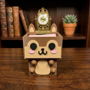 Puede incluir: Un organizador de escritorio marrón y beige impreso en 3D con forma de ardilla de dibujos animados. El organizador tiene forma de caja rectangular con una cara de ardilla en la parte delantera, que incluye ojos negros, mejillas rosas y una boca negra pequeña. Las orejas son beige y marrón.