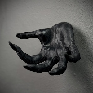 May include: A matte black wall-mounted sculpture of a hand with elongated, claw-like fingers. The surface features detailed, realistic skin textures, including visible wrinkles and knuckles, extending from the wall in a grasping, open-palm gesture.