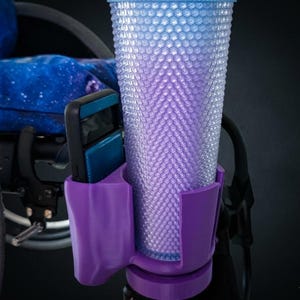 Peut inclure: Un porte-gobelet et porte-téléphone en plastique violet fixé sur le cadre d'un fauteuil roulant. Le support contient un grand gobelet texturé dégradé bleu clair à violet avec un motif géométrique et un smartphone sombre avec une coque bleue.