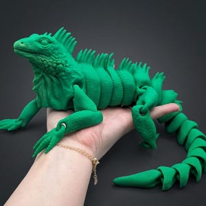 Puede incluir: Una figura de iguana articulada de color verde brillante con una superficie texturizada y escamosa, una cresta dorsal espinosa a lo largo de la espalda y una cola segmentada y flexible. El lagarto tiene articulaciones móviles en extremidades.