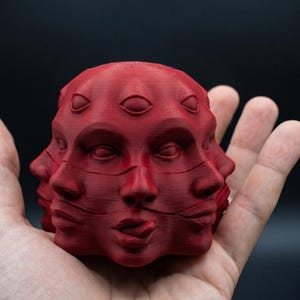 May include: A matte red 3D printed sculpture featuring multiple human faces merged into a single spherical form. The object displays several eyes and mouths arranged around the surface, with one mouth slightly open showing a tongue, all in a solid red.