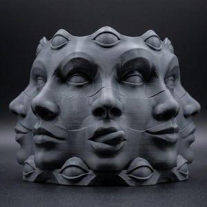 Peut inclure: Un pot de fleurs cylindrique gris mat présentant un motif répétitif de visages humains sur tout son extérieur. Chaque visage affiche des traits distincts incluant yeux, nez et lèvres, avec un visage montrant une langue tirée. Les bords supérieur et inférieur présentent une série de motifs d'yeux sculptés.
