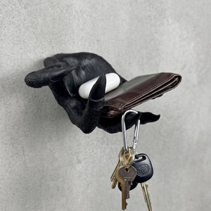 May include: A wall-mounted black claw-shaped hand sculpture holding a brown leather wallet and a white wireless earbud case. A silver carabiner with a set of metal keys, including a black Toyota car key fob, hangs from the fingers of the claw.