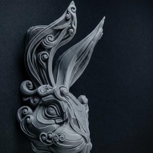 Può includere: Una scultura da parete grigio opaco a forma di testa di coniglio. Il design presenta intricati motivi in rilievo, vorticosi e simili a nuvole, integrati nelle orecchie e nei tratti del viso, creando un aspetto strutturato e stratificato.