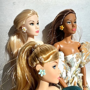 May include: Three fashion dolls with blonde and brown hair. Each doll wears flower-shaped earrings in white, yellow, and light blue. One doll wears a floral print dress. The dolls have detailed facial features, including blue eyes and pink lips.