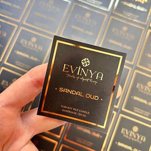 以下が含まれることがあります： 手で持たれた、黒と金のキャンドルのラベル。ラベルには「EVINYA」と「Sandal Oud」と書かれています。また、キャンドルは100％大豆ワックス、手作り、250 mlであることも示しています。背景には他の同様のラベルがあります。