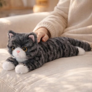 Puede incluir: Un peluche de gato atigrado gris y negro, con patas blancas y nariz rosa. El gato está tumbado sobre una superficie beige. Tiene ojos grandes y redondos amarillos y orejas rosas.