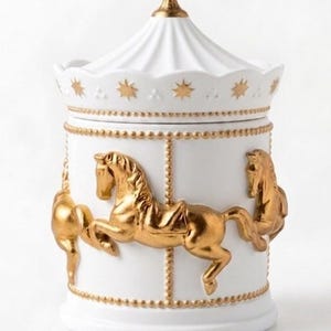 May include: White ceramic carousel jar with gold detailing. The jar features gold carousel horses, star details, and a gold-tipped lid. The jar is a decorative storage container.