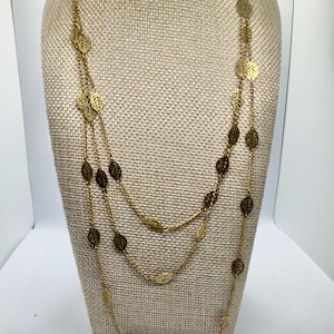 May include: A gold-toned necklace with three strands, each adorned with delicate leaf-shaped charms. The necklace is displayed on a neutral-toned mannequin, showcasing its layered design and intricate details. The leaves have a detailed cut-out design.