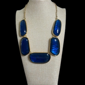 May include: A statement necklace featuring large, irregular blue stones set in gold-tone metal. The necklace is displayed on a mannequin, showcasing its bold design and color. The stones have a glossy finish.