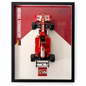 Peut inclure: Un cadre présentant un modèle réduit de voiture de Formule 1 rouge et blanche. Le fond inclut le texte MICHAEL SCHUMACHER, un drapeau allemand, une signature, ainsi que les logos Marlboro et Ferrari. La voiture est placée verticalement.