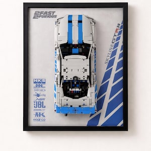 May include: Framed model car of a white and blue Skyline GT-R34, a replica from the movie 2 Fast 2 Furious. The model has blue racing stripes and sponsor logos. The frame is black.