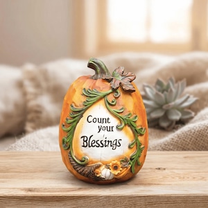 Könnte beinhalten: Orangefarbene Kürbis-Dekoration mit dem Text "Count your Blessings". Das Objekt hat grüne Ranken- und Blätterverzierungen, einen braunen Stiel und ein kleines Füllhorn- und Sonnenblumen-Design. Es steht auf einer Holzoberfläche.