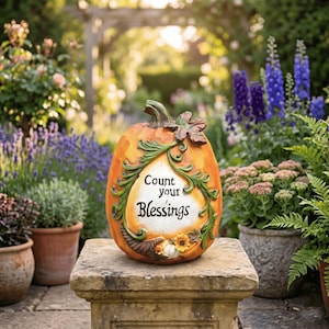 Könnte beinhalten: Eine orangefarbene Kürbis-Gartenstatue mit dem Schriftzug "Count your Blessings" in einem weißen Oval. Die Statue hat grüne Blatt- und Rankenverzierungen und steht auf einem Steinsockel in einem Garten.