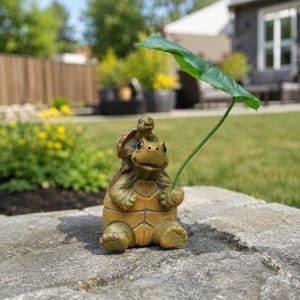 May include: A decorative garden figurine featuring a large brown turtle sitting upright with a smaller turtle perched on its back. The large turtle holds a single long green stem with a large, broad green leaf extending upward and outward.