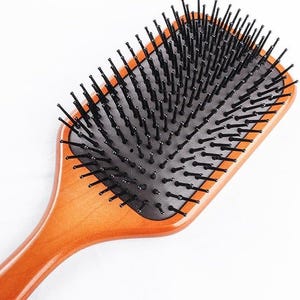 May include: A rectangular hairbrush with a smooth, light brown wooden handle and base. The brush features a black rubber cushion embedded with numerous black plastic bristles, each topped with a small, rounded tip for detangling hair.