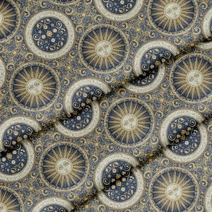 Op de afbeelding: Stof met een herhalend hemels patroon met zonnen, manen en sterren in goud, marineblauw en crème. Het ontwerp bevat sierlijke krullen en geometrische vormen, wat een vintage of bohemien esthetiek creëert. Geschikt voor kleding of woondecoratie.