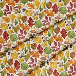 May include: A fabric featuring a repeating pattern of autumn leaves in shades of yellow, orange, red, and green, interspersed with small brown acorns and spiral-shaped seed pods on a light cream-colored background, shown with a diagonal fold.