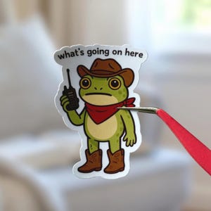 Puede incluir: Una pegatina con un dibujo de una rana vestida de vaquero, con sombrero marrón, pañuelo rojo y botas. La rana sostiene un walkie-talkie y el texto "what's going on here" está encima.