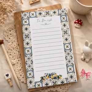Può includere: Un blocco note rettangolare con un centro bianco caratterizzato da linee orizzontali e dal testo To-Do-ish con un piccolo cuore. Il bordo è costituito da un motivo a piastrelle blu, giallo e bianco, con un grappolo di limoni e foglie in basso.
