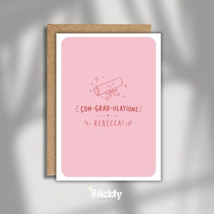 Peut inclure: Une carte de vœux rose clair avec une enveloppe brune. La carte présente un dessin au trait d'un diplôme noué avec un ruban et le texte CON-GRAD-ULATIONS REBECCA! en encre rouge. Le design inclut de petites étoiles et des feuilles.