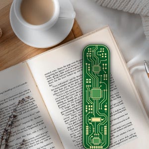 Puede incluir: Un marcapáginas verde con forma de placa de circuito impreso descansa sobre un libro abierto, junto a una taza de café. El marcapáginas presenta un diseño detallado de circuitos y componentes electrónicos.