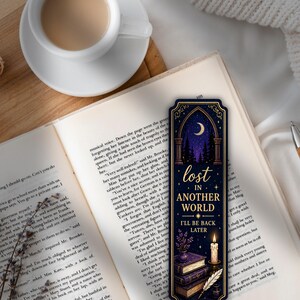 May include: A bookmark with a night sky design, a crescent moon, and the words "lost in another world, I'll be back later." It rests on an open book, beside a cup of coffee. The bookmark has a decorative border.