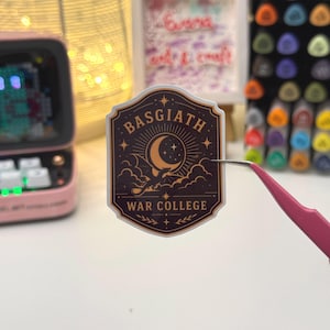 May include: A brown and gold sticker featuring the text "BASGIATH WAR COLLEGE" and a crescent moon design. The sticker is held by pink tweezers. The sticker has a shield shape with a dragon and star design.