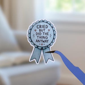 Peut inclure: Un autocollant en forme de ruban de récompense bleu clair avec deux pans, portant le texte noir "CRIED BUT DID THE THING ANYWAY" et de petites illustrations de cœurs roses. L'autocollant est tenu par une pince bleue sur un fond flou.