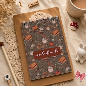 Può includere: Un taccuino a spirale con una copertina grigio scuro caratterizzata da un motivo ripetuto di zucche, tazze, libri e sciarpe. Un'etichetta rettangolare color ruggine al centro mostra la parola notebook in corsivo bianco.