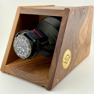 May include: A wooden watch winder with a black watch featuring a black dial and a blue and red strap. The winder is made of dark wood and has a gold-colored emblem on the side.