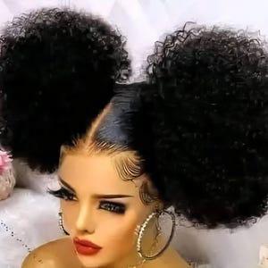 Può includere: Una testa di manichino con una parrucca nera caratterizzata da due grandi e voluminosi chignon afro su entrambi i lati. I capelli sono nero scuro con ricci stretti e il manichino presenta bordi definiti, lunghe ciglia scure e labbra rosse.
