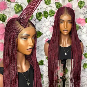 May include: Two wigs with long, burgundy braids are displayed. The wigs are on mannequin heads, showcasing the intricate braiding pattern and the natural-looking hairline. The background features a floral and leaf pattern.