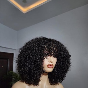 Könnte beinhalten: Eine schwarze, lockige Perücke mit Pony wird auf einem Schaufensterkopf präsentiert. Die Perücke hat einen vollen, voluminösen Stil mit engen Locken. Die Schaufensterpuppe hat einen neutralen Hautton und steht vor einem neutralen Hintergrund.