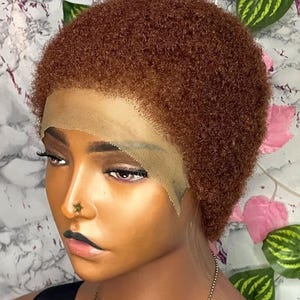 Op de afbeelding: Een korte, roodbruine pruik met een kanten voorkant wordt getoond op een mannequin. De pruik heeft een strak gekrulde textuur en is dicht op het hoofd gestyled. De mannequin heeft donkere make-up en een gouden neusring. De achtergrond heeft roze bloemen en groene bladeren.