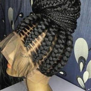 Puede incluir: Una peluca trenzada negra con una parte delantera de encaje se muestra en una cabeza de maniquí. Las trenzas están tejidas apretadamente y peinadas en un moño. La parte delantera de encaje es de tono de piel claro. El fondo es azul oscuro con un patrón floral blanco y negro.