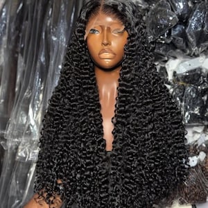 May include: A black curly wig on a mannequin head. The wig has a realistic hairline and long, voluminous curls. The mannequin has brown skin. The background shows packaged items.
