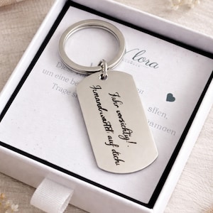 Könnte beinhalten: Ein silberfarbener Metall-Schlüsselanhänger mit einem rechteckigen Anhänger, präsentiert in einer weißen Geschenkbox. Der Anhänger trägt den eingravierten Text "Fahr vorsichtig! Jemand wartet auf dich." Der Schlüsselanhänger ist an einem großen Ring befestigt.