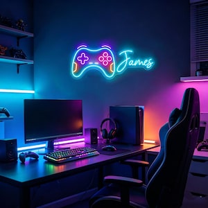 Op de afbeelding: Een neonbord in de vorm van een gamecontroller met de naam "James" in een scriptlettertype. Het bord is verlicht met blauwe, oranje en roze lichten. Het is gemonteerd op een donkerblauwe muur boven een gaming-opstelling met een monitor, toetsenbord en gamestoel.