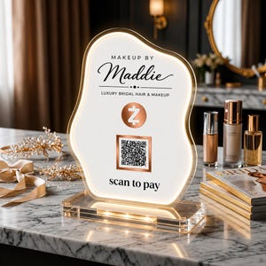 May include: An illuminated, organic-shaped acrylic sign featuring the text "MAKEUP BY Maddie" and "LUXURY BRIDAL HAIR & MAKEUP." It includes a Zelle logo, a square QR code, and the text "scan to pay" printed on a white surface with a gold border.