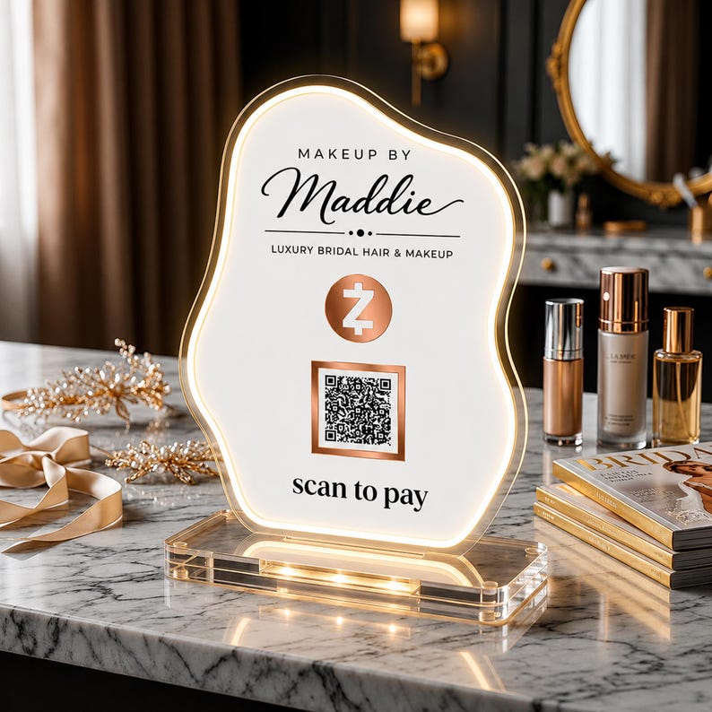 May include: An illuminated, organic-shaped acrylic sign featuring the text "MAKEUP BY Maddie" and "LUXURY BRIDAL HAIR & MAKEUP." It includes a Zelle logo, a square QR code, and the text "scan to pay" printed on a white surface with a gold border.