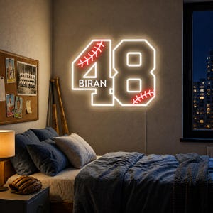 May include: A wall-mounted neon sign shaped like the number 48, featuring white illumination and red baseball stitching details. The name BIRAN is displayed in white capital letters within the number four. The sign emits a bright glow.