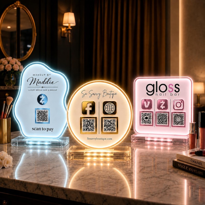 May include: Three illuminated acrylic signs on a marble surface. The left sign is blue with 'Makeup by Maddie' and a QR code. The center sign is yellow with 'So Savvy Boutique' and social icons. The right sign is pink with 'gloss nail bar' and QR codes.