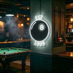 May include: A neon sign in the shape of an eight ball, with the text "EST 1999". The sign is white with a black eight ball in the center. A pool table and cue sticks are visible in the background.
