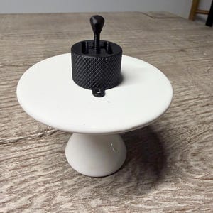May include: A black 3D printed gear shifter knob featuring a textured knurled grip pattern on the cylindrical base and a central vertical stick. The object is positioned on a smooth white circular pedestal stand with a flared base.