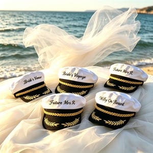 Może przedstawiać: Pięć białych czapek kapitańskich z czarnymi daszkami i złotymi detalami. Na każdej czapce napis: "Bride's Voyage", "Future Mrs R" i "Wifey On Board". Czapki mają złote sznurkowe wykończenia i złote wzory liści, na tle plaży.