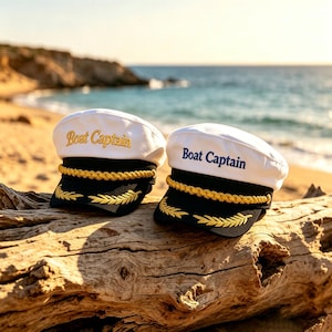 May include: Two white boat captain hats with black visors and gold accents rest on a piece of driftwood. The hats have the words "Boat Captain" embroidered on the front. The background shows a beach and the ocean.