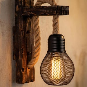 May include: Rustic wall sconce featuring a wooden base and a woven rope detail. The light fixture has a black metal cage shade and a warm, glowing bulb. The design evokes a vintage, industrial aesthetic, perfect for adding character to any space.