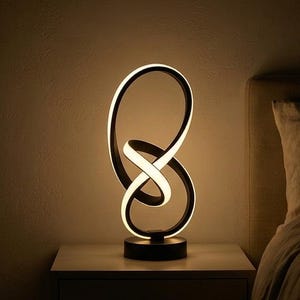 Op de afbeelding: Een moderne zwart-witte LED-tafellamp met een uniek, verweven lusontwerp. De lamp heeft een ronde voet en straalt een warm licht uit. Hij staat op een wit nachtkastje naast een bed.