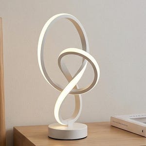 Op de afbeelding: Een moderne witte LED-tafellamp met een uniek, verweven lusontwerp. De lamp heeft een cirkelvormige basis en een vloeiende, verlichte vorm, waardoor een zachte, omgevingsgloed ontstaat. De lamp staat op een houten oppervlak.